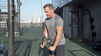 Un hombre caucásico de mediana edad, vestido con camiseta gris y pantalones cortos oscuros, realiza un pushdown de tríceps con una cuerda en un gimnasio al aire libre, con una ciudad al fondo.
