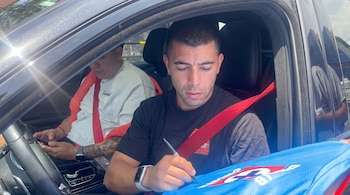 Un hombre con camiseta negra firma un objeto azul desde la ventana de un vehículo, mientras otra persona permanece sentada al lado.