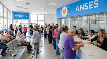 Interior de una oficina de ANSES con personas haciendo fila y sentadas, mientras empleados atienden en mostradores, algunos entregando dinero. Hay carteles de ANSES.