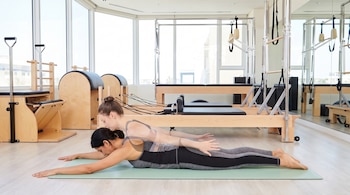Dos mujeres ejercitándose en un estudio de pilates con máquinas de madera y metal. Una está acostada en una colchoneta, la otra se muestra en movimiento translúcido.