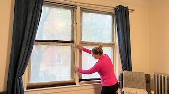 Mujer con camiseta rosa aplicando una película de plástico transparente a una ventana, con cortinas azules y burletes en el alféizar, para aislar del frío.