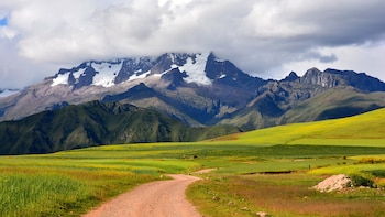 Un camino de tierra atraviesa campos de cultivo verdes y amarillos hacia montañas con picos nevados parcialmente cubiertos de nubes