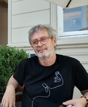 Primer plano de un hombre mayor con cabello y barba grises, gafas de montura marrón y una camiseta negra con un dibujo lineal blanco, sentado al aire libre