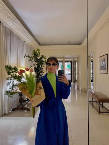 Una mujer con abrigo azul y gafas de sol púrpuras sostiene un ramo de tulipanes rojos y un teléfono móvil, reflejándose en un espejo