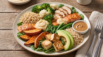 Plato circular sobre mesa de madera que contiene porciones de salmón, pollo, tofu, verduras, arroz, quinoa, batata, palta y salsa.