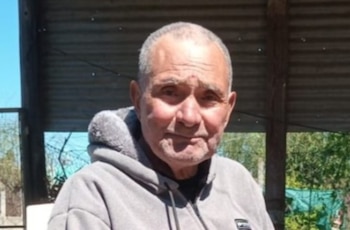 Hombre mayor con cabello gris y calvicie frontal, viste buzo gris con capucha y cremallera, posando al aire libre