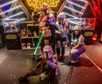Snarky Jay arrodillada y sonriente junto a tres cosplayers de Mandalorian con sables de luz y Grogu, en un evento temático de Star Wars