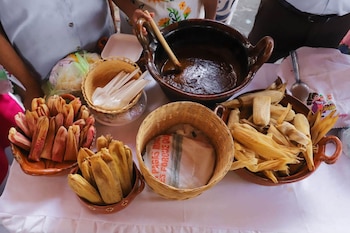 Mesa con recipientes de barro y cestas con tamales de hojas de maíz, salsa oscura en olla de barro, cucharas plásticas, y tela blanca