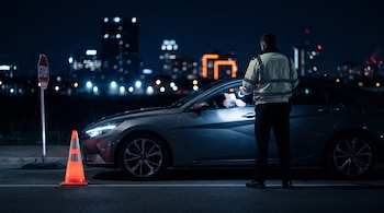Agente de tránsito de espaldas con chaleco reflectante ilumina documentos de conductor en auto gris detenido, cono naranja y señal de STOP en la calle, ciudad de noche.