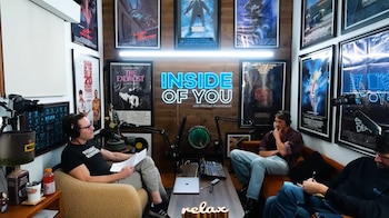 Dos hombres, Michael Rosenbaum y Joel Kinnaman, con auriculares en un estudio de podcast, frente a un letrero de neón y carteles de películas en la pared