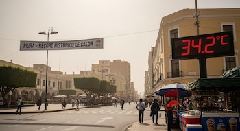 Calle con personas, edificios, un termómetro digital que muestra 34.2 °C, un cartel con "PIURA - RÉCORD HISTÓRICO DE CALOR" y puestos.