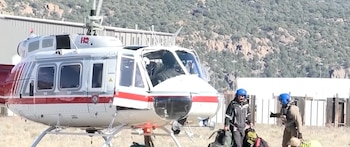 Un helicóptero blanco y rojo posado en un terreno despejado junto a dos personas con cascos azules que cargan mochilas y equipo de trabajo