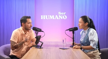 Katy Jara y un hombre sentados en una mesa de madera con micrófonos de podcast, frente a un fondo morado con el texto "Ser HUMANO"