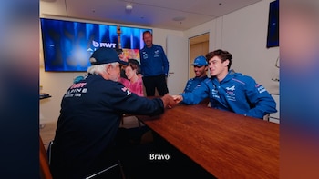 Cuatro integrantes del equipo Alpine de Fórmula 1 con ropa deportiva azul conversan y se dan la mano alrededor de una mesa de madera