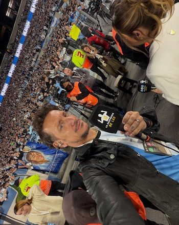Nico Vázquez sostiene un micrófono frente a la cámara en un estadio rodeado de público y personal técnico