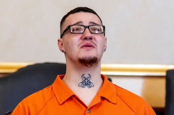 Primer plano de un hombre joven con gafas, perilla y un tatuaje de polilla en el cuello, vistiendo un uniforme naranja. Tiene lágrimas en los ojos