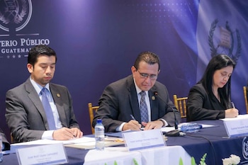Tres personas con vestimenta formal sentadas a una mesa firmando documentos frente al escudo del Ministerio Público de Guatemala