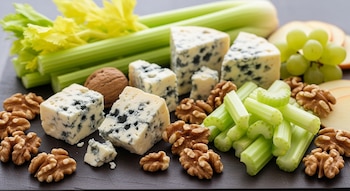 Varias porciones de queso Roquefort, apio fresco en tallos y trozos, y nueces peladas sobre una tabla oscura, junto a uvas verdes y rodajas de manzana.