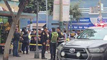 Agentes de policía y un vehículo policial con luces encendidas acordonan una zona urbana con cinta amarilla y personas observando