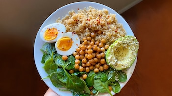 Primer plano de un plato blanco con quinoa, garbanzos asados, hojas de espinaca, dos mitades de huevo cocido y medio aguacate con semillas de sésamo.