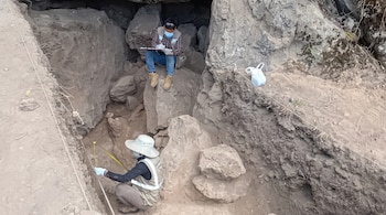 Dos personas con mascarillas en una excavación arqueológica miden una trinchera de tierra y rocas; la otra persona registra datos sentada entre rocas