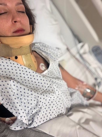 Mujer joven acostada en cama de hospital, con ojos cerrados y collarín cervical beige. Viste camisón hospitalario y tiene catéter intravenoso en el brazo