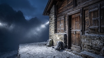 Fachada de un refugio de piedra y madera cerrado con candado, dos mochilas cubiertas de nieve en el suelo y montañas de fondo de noche.