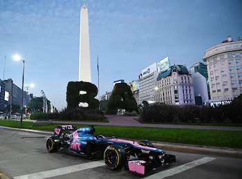 Monoplaza de Fórmula 1 rosa y azul oscuro en una calle de Buenos Aires, con el Obelisco y letras "BA" vegetales al fondo, bajo un cielo crepuscular