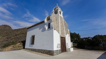 Ermita blanca con campanario, cruz, campana, fachada de piedra. Puerta de madera, ventana con reja. Terreno de cemento, colina, árboles, cielo azul