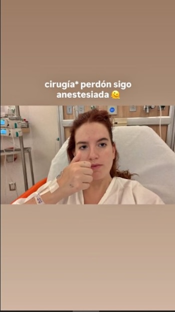 Nicole Zignago en cama de hospital, vestida con bata blanca, brazo izquierdo con vía intravenosa, haciendo un pulgar arriba. Tiene el cabello pelirrojo