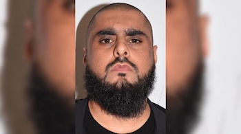 Un hombre de piel oscura con cabeza rapada, barba y bigote negros, cejas gruesas y ojos marrones, sobre un fondo liso y claro.