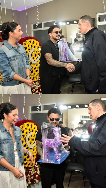 Un hombre tatuado con gafas de sol y una mujer con chaqueta de mezclilla observan a otro hombre entregar un alebrije morado en un evento