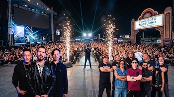 Integrantes de grupos musicales en primer plano ante una multitud en un concierto nocturno con luces y pirotecnia.