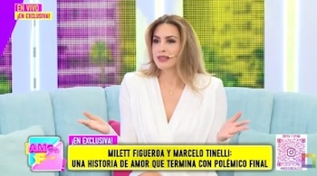 Milett Figueroa sentada en un sofá azul claro en un set de televisión, vistiendo un vestido blanco y con un micrófono. Tiene las manos abiertas mientras habla