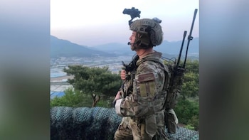 Un militar con casco, antenas de radio en la espalda y uniforme de camuflaje frente a un valle y colinas al fondo