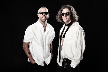 Dos hombres usan gafas de sol y camisas blancas con pantalones oscuros. Uno lleva cadena plateada y el otro, corbata negra sobre un fondo negro