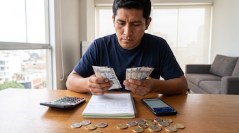 Hombre revisa billetes de soles y monedas sobre una mesa, junto a una calculadora, un cuaderno con anotaciones y un teléfono móvil con aplicación bancaria.