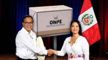 Dois sorridentes candidatos presidenciais, um homem e uma mulher, de mãos dadas diante da ânfora do ONPE e da bandeira do Peru, no pódio.