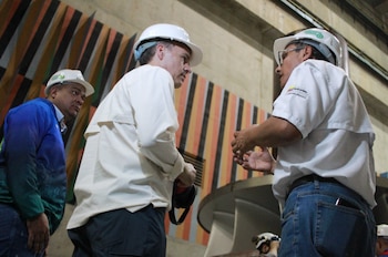 Tres hombres con cascos de seguridad blancos dentro de una instalación industrial. Dos conversan de perfil. Una pared con franjas verticales de colores
