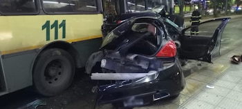 Primer plano de un autobús amarillo con el número 111, un automóvil negro chocado y un bombero. Se observan restos del vehículo
