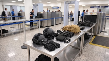 Cascos tácticos, gafas protectoras, guantes, chalecos y mochilas sobre una mesa metálica frente a escáneres e inspectores en un aeropuerto.