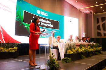 Mujer de cabello oscuro con vestido rojo habla desde podio transparente. Detrás, una pantalla grande con logo Gobierno de México y texto 'Calificación e Inversión'