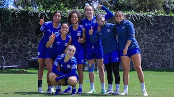 Siete mujeres con uniformes deportivos azules posan en un campo de césped verde; una está agachada, con una pared de piedra y vegetación al fondo.
