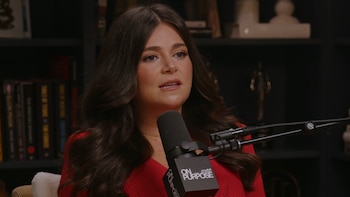 Una mujer de cabello castaño oscuro y suéter rojo habla frente a un micrófono con el logo de "On Purpose by Jay Shetty", con estanterías borrosas detrás