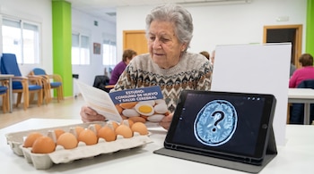 Primer plano de una mujer mayor con canas leyendo un folleto. Sobre la mesa hay un cartón de huevos y una tableta mostrando un cerebro con interrogación.