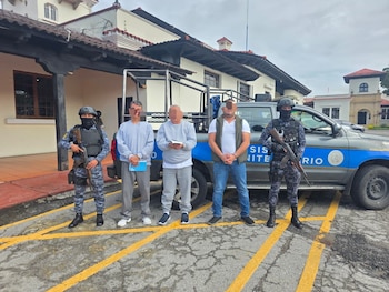 Un agente armado custodia a cuatro hombres esposados con rostros difuminados, parados en hilera frente a un cartel y una ventana