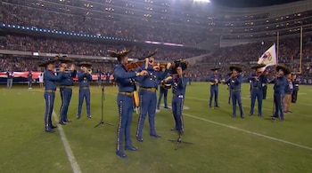 Músicos vestidos de mariachi azul tocan violines y trompetas en el césped de un estadio deportivo durante la noche.