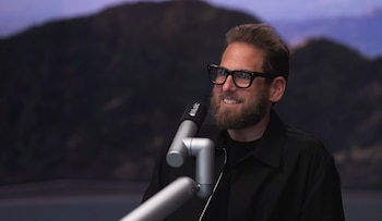 Un hombre con barba y gafas, identificado como Jonah Hill, sonríe ampliamente mientras está sentado frente a un micrófono de Apple Music. El fondo es un paisaje montañoso borroso