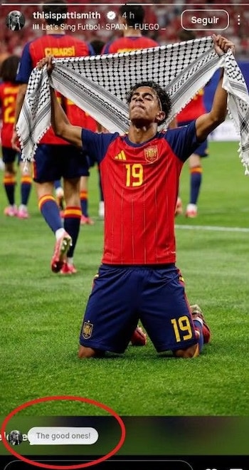 Jugador Lamine Yamal arrodillado en campo de fútbol, con camiseta roja número 19, shorts azules, sosteniendo una keffiyeh blanca y negra. Otros jugadores en el fondo