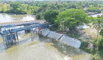 Vista aérea de un puente de concreto sobre un río turbio con tuberías azules, con muros de contención en construcción en una orilla y vegetación densa